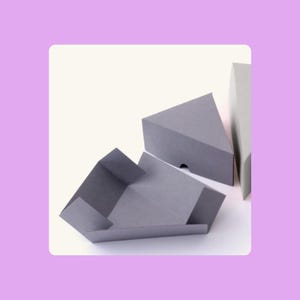 Puede incluir: Tres cajas triangulares grises sobre un fondo blanco. Una caja está abierta, revelando su interior, las otras están cerradas. Las cajas están hechas de un material mate.