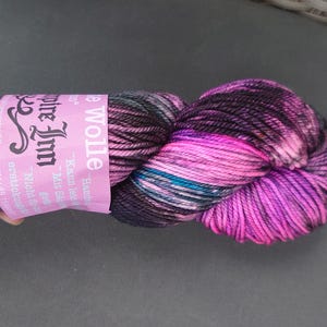 Könnte beinhalten: Ein Strang handgefärbter Wolle in Rosa-, Lila-, Schwarz- und Türkistönen. Die Wolle ist mit einem rosa Etikett mit deutschem Text umwickelt. Die Wolle wird in einer Hand gehalten.
