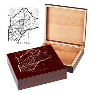 May include: A wooden box with a hinged lid. The top of the box is a dark red color and features a gold-colored map of Malaga, Spain. The lid is made of light wood and is open to reveal the interior of the box.