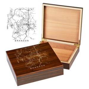 May include: A wooden box with a hinged lid, featuring a detailed map of Branson, Missouri, engraved on the top. The box is open, revealing a light-colored interior. The text "BRANSON" is visible on the box.