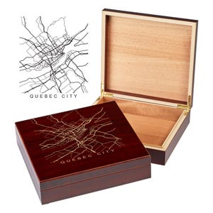 May include: A wooden box with a hinged lid. The top of the box is a dark red color and features a gold-colored outline of the city of Quebec. The lid is made of light wood and is open to reveal the interior of the box.