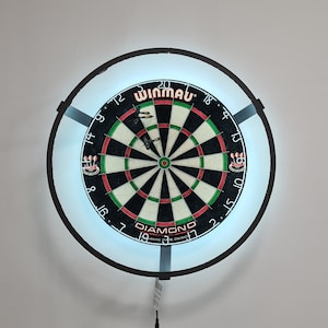 May include: A black dartboard with a white bullseye and red and green rings. The dartboard is surrounded by a black ring with a white halo. The dartboard is mounted on a white wall. The brand name "Winmau" is printed on the dartboard.