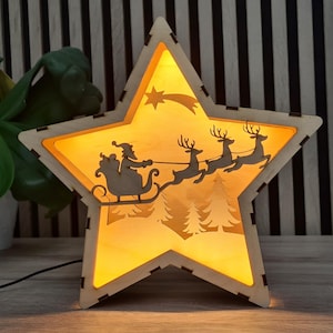 May include: Wooden star-shaped Christmas decoration with a warm yellow glow. The star features a silhouette of Santa Claus in a sleigh pulled by reindeer, with a shooting star and pine trees. A festive holiday accent.