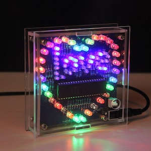 Könnte beinhalten: Eine quadratische digitale Uhr aus klarem Acryl mit einem schwarzen Leiterplatten-Innenraum. Die Uhr zeigt die Zeit mit farbigen LED-Leuchten in einem kreisförmigen Muster an. Die Lichter sind in verschiedenen Farben, darunter Rot, Grün und Blau.