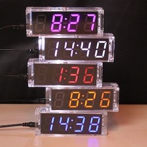 Könnte beinhalten: Fünf transparente Plastikgehäuse, die übereinander gestapelt sind, jedes mit einer digitalen Uhr mit unterschiedlich farbigen LED-Anzeigen. Die Uhren zeigen die Zeiten 8:27, 14:40, 1:36, 8:26 und 14:38 an.