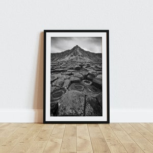 May include: Black and white framed photograph of a natural landscape. The image features a unique rock formation in the foreground, leading to a mountain in the background. The photograph is displayed on a wooden floor.