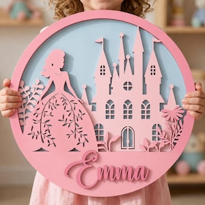 May include: A circular wooden wall sign featuring a light blue background with a layered pink cutout design. The design includes a silhouette of a figure in a gown, a castle with multiple towers, floral accents, and the name Emma in script font.