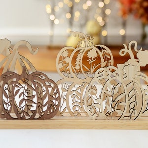 May include: Three wooden pumpkins with intricate cut-out designs of leaves and flowers. The pumpkins are arranged on a wooden base.