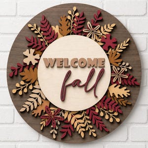 May include: A circular wooden wall sign featuring a dark brown base layered with cutout leaves in shades of burgundy, tan, and cream. The center displays a light wood circle with the text WELCOME fall in raised, dark brown lettering.