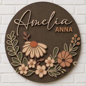 Könnte beinhalten: Runder Holzschild mit dem Namen "Amelia" in Schreibschrift und dem Namen "Anna" in Blockbuchstaben. Das Schild ist mit einem Blumendesign in Beige-, Orange- und Grüntönen verziert. Der Hintergrund ist dunkelbraun.