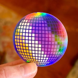 May include: A round, holographic sticker featuring a disco ball design. The sticker has a grid pattern with a gradient of colors, including purple, yellow, and green. The sticker is held between two fingers.
