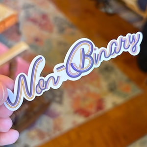 May include: A white sticker with the words "Non-Binary" in a stylized font. The text is outlined in purple and gold, with a purple and gold gradient within the letters. The sticker is held against a blurred background.