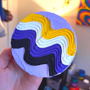 May include: A round, lavender-colored art piece with a textured surface. The design features wavy bands of black, purple, white, and yellow. The piece is held in a hand.