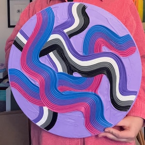 Op de afbeelding: Een rond, getextureerd kunstwerk met een lavendelkleurige achtergrond. Golvende, verhoogde lijnen in tinten blauw, roze, paars, grijs, zwart en wit creëren een dynamisch, abstract ontwerp. Het stuk wordt vastgehouden door iemand met een roze jas.