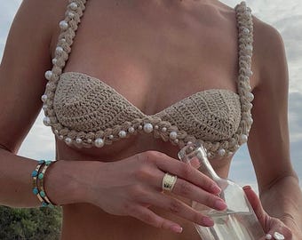 Puede incluir: Un top y una parte inferior de bikini de crochet con detalles de perlas. El bikini es beige y las perlas son blancas. La modelo sostiene una botella de vidrio transparente.