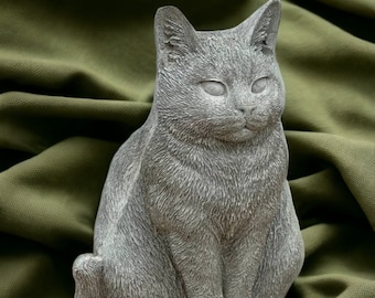 May include: A gray stone cat figurine sitting on a green fabric background. The cat is sitting with its front paws tucked under its body and its head tilted slightly to the side.