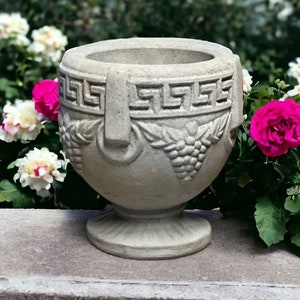May include: A gray stone planter with a Greek key design and grapevine accents. The planter is sitting on a stone pedestal surrounded by white and pink roses.