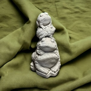 May include: A stack of three gray ceramic frog figurines. The frogs are stacked on top of each other, with the largest frog at the bottom and the smallest frog at the top. The frogs are all facing forward and have their eyes closed.