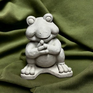 May include: A gray concrete frog figurine with a wide smile and clasped hands. The frog is sitting on a green fabric background.