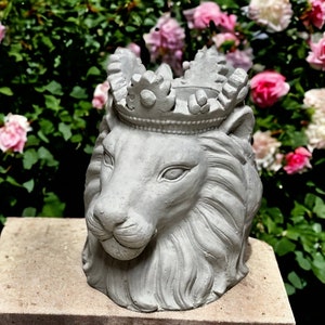 May include: A gray concrete lion head planter with a crown on top. The planter is sitting on a gray stone surface. The background is a blurred image of pink and white flowers.