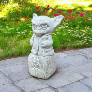 以下が含まれることがあります： スターウォーズのヨーダの白いコンクリート像が、ダース・ベイダーの彫刻が施された台座の上に立っています。像は石畳の屋外にあります。