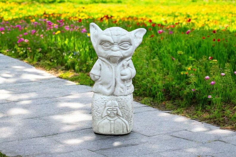 以下が含まれることがあります： スターウォーズのヨーダの白いコンクリート製の庭の像で、ダース・ベイダーを描いた台座の上に立っています。像は庭の石畳の上にあります。