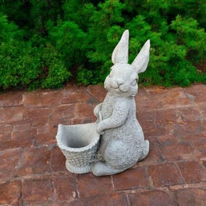 May include: A gray concrete rabbit statue holding a basket planter. The rabbit has a whimsical expression and is standing on a brick patio.