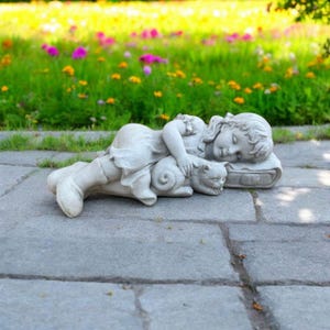 May include: A gray stone statue of a sleeping child with a cat. The child is lying down, resting their head on a pillow, and cuddling the cat. The statue is set on a stone surface with a background of green grass and colorful flowers.