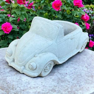 May include: A white ceramic planter shaped like a classic Volkswagen Beetle convertible. The planter has a rounded shape and a detailed design, including headlights, a windshield, and a removable top for planting.
