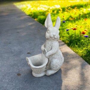 May include: A light gray bunny statue holding a basket. The bunny has long ears and a smiling face. The basket is empty and ready to be filled. The statue is made of a stone-like material and is suitable for outdoor use.