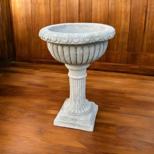 May include: A gray, ornate pedestal planter with a wide, scalloped rim. The planter has a fluted body and a square base. The overall height is approximately 30 inches.