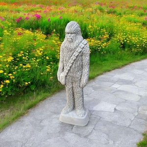 May include: A gray stone statue of a Wookiee standing on a stone base. The statue is in a garden setting with a stone path leading away from it.