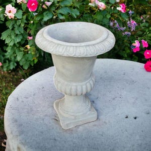 May include: A white concrete urn with a decorative rim and a fluted base. The urn is sitting on a gray concrete surface.