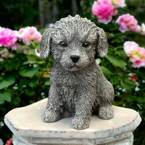 May include: A gray, textured dog statue with a curly coat, sitting on a stone pedestal. The dog has a black nose and large, round eyes. The background features pink flowers and green foliage.