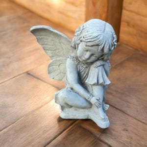 May include: A light gray stone-colored statue of a seated figure with large, detailed wings folded behind the back. The figure has short, textured hair, a simple dress, and is posed with one arm resting on a knee, looking downward in a quiet manner.