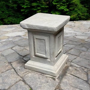 May include: A white concrete pedestal with a square top and decorative molding. The pedestal is sitting on a stone patio.