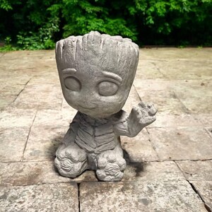 May include: A gray concrete planter shaped like a cartoon character with large eyes and a textured surface. The planter is in a seated position with one hand raised. The design is detailed, with visible facial features and body textures.
