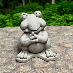 May include: A gray concrete frog statue with a wide smile and its hands clasped together in front of its chest. The frog is sitting on a gray stone surface.