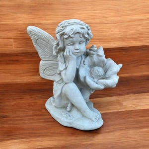 May include: A gray concrete fairy statue with detailed wings, holding a large flower. The fairy is seated, resting her chin on her hand, with a serene expression. The statue is on a wooden surface.