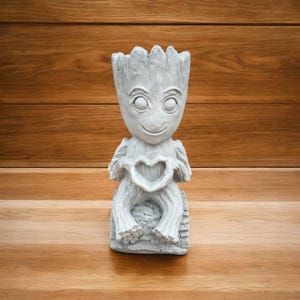 May include: A light gray concrete planter shaped like a small tree-like creature with large eyes and a textured bark-like surface. The figure is seated with its hands positioned in front of its chest to form a heart-shaped opening in the center.