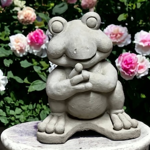 May include: A gray concrete frog statue with a wide smile and its hands clasped together. The frog is sitting on a round pedestal in front of a background of pink and white roses.