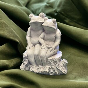 May include: A pair of gray ceramic frogs sitting on a tree stump. The frogs are holding each other and have a sweet expression on their faces.