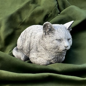 May include: A gray, sleeping cat figurine made of concrete or stone. The figurine is lying on a green fabric surface.