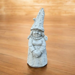 May include: A gray concrete garden gnome statue. The gnome has a tall, textured hat and is wearing a dress. The gnome is smiling and has its hands clasped in front of it. The statue is on a wooden surface.