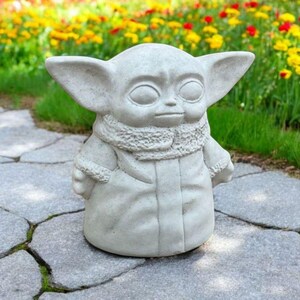 Pu&ograve; includere: Una statua di cemento grigio di un personaggio del franchise di Star Wars, noto come "The Child" o "Baby Yoda". La statua &egrave; in piedi su un sentiero di pietra con un'aiuola di fiori sullo sfondo.