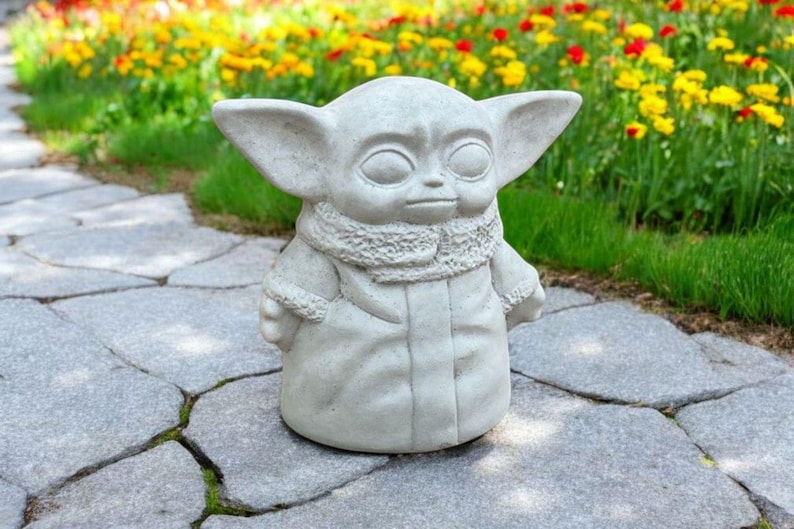 Pu&ograve; includere: Una statua di cemento grigio di un personaggio del franchise di Star Wars, noto come "The Child" o "Baby Yoda". La statua &egrave; in piedi su un sentiero di pietra con un'aiuola di fiori sullo sfondo.