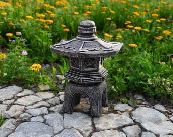 May include: A gray stone pagoda lantern with a detailed design. The lantern is sitting on a stone path in a garden setting.