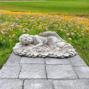 May include: A white stone sculpture of a fairy lying on a bed of leaves. The sculpture is placed on a stone patio with a field of flowers in the background.