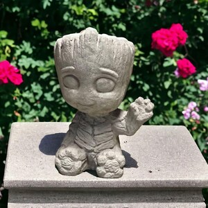 May include: A gray concrete garden statue of a character from the movie Guardians of the Galaxy. The statue is sitting on a gray concrete pedestal. The statue is in a garden setting with pink and red flowers in the background.