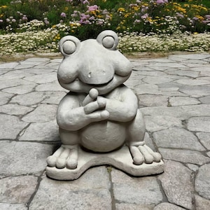 May include: A gray stone frog statue with a wide smile and its hands clasped together. The frog is sitting on a stone base.